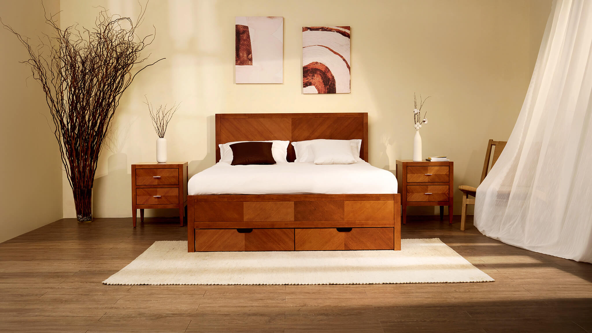 "A frontal view of a modern bedroom scene centered on a large wooden storage bed with a geometric inlay pattern on its headboard and footboard. The footboard features two integrated storage drawers. Matching three-drawer nightstands flank the bed. White linens and a mix of white and one dark brown pillow are on the bed. A tall dried branch arrangement is in a large vase to the far left. Smaller vases with branch and cotton stems are on the nightstands. Two abstract organic art prints in brown and rust tones