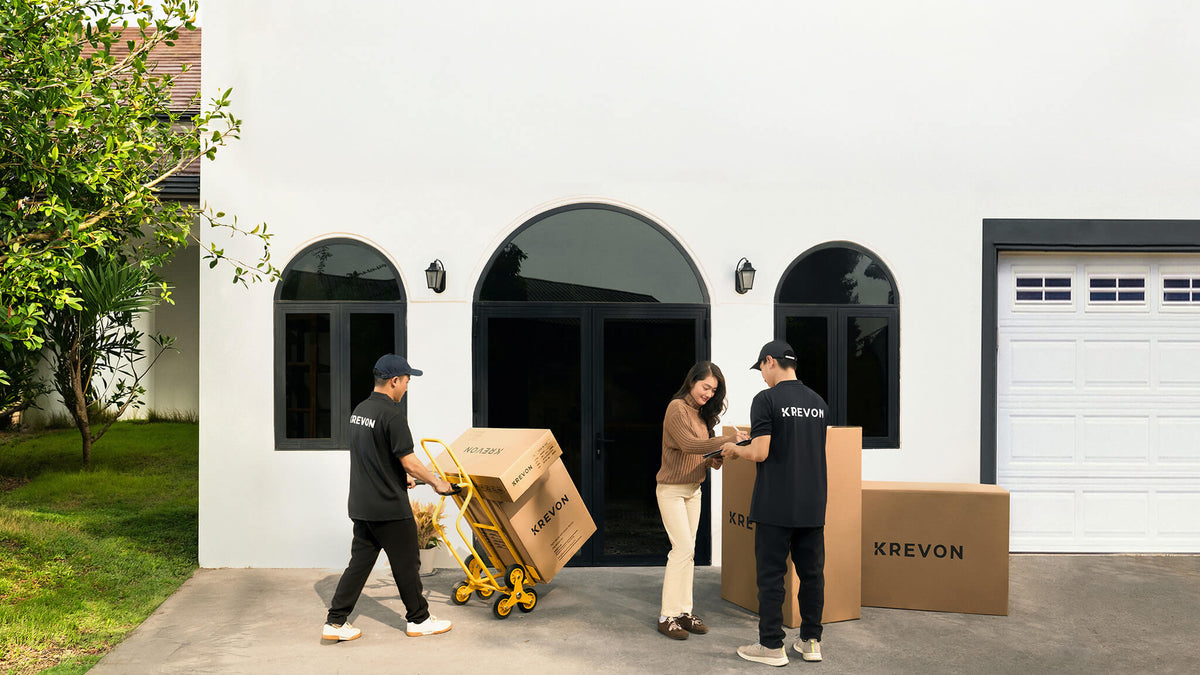 Two KREVON furniture delivery personnel, wearing black uniforms with 'KREVON' printed on them and matching hats, are seen outside a modern white building with arched windows. One delivery person on the left pushes a yellow trolley with multiple KREVON-branded boxes. The other delivery person on the right is with a female customer with long dark hair, wearing a brown sweater and light-colored pants. He is holding a tablet, and they are reviewing the delivery details as the customer stands next to large KREVO